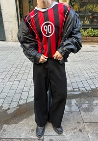 Maillot Nike rayé verticalement rouge et noir avec un graphique circulaire "90", associé à un pantalon noir ample et des chaussures noires, et une doudoune oversize.