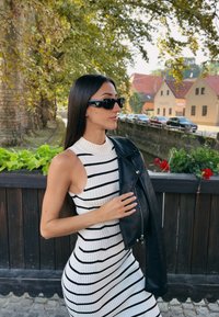Černá kožená bunda přes bílé a černé pruhované žebrované šaty. Sluneční brýle s logem. Pozadí zahrnuje zeleň a rustikální budovy.