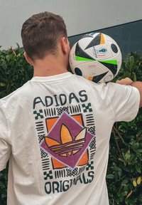 Λευκό κοντομάνικο μπλουζάκι από βαμβάκι με πολύχρωμο γραφικό σχέδιο που περιλαμβάνει γεωμετρικά σχήματα και το κείμενο "ADIDAS ORIGINALS" στην πλάτη.