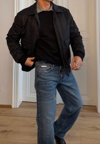 Czarna kurtka bombowiec o gładkiej fakturze, noszona na ciemnym swetrze. Jasnoniebieskie, przetarte dżinsy z logiem Calvin Klein oraz srebrny pierścionek.