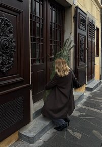 Personne portant un manteau marron et des talons hauts noirs, tenant un arbre vert devant des portes doubles en bois avec des panneaux en verre et des éléments décoratifs.