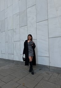Černý kabát přes úzké, třpytivé šedé šaty, černé kozačky po kolena a malá černá kabelka. Hladká, texturovaná zeď v pozadí.