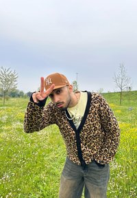 Καρφίτσα με λεοπάρ σχέδιο και μαύρες λεπτομέρειες, φορεμένη πάνω από ένα ανοιχτόχρωμο μπλουζάκι με σχέδιο και συνδυασμένη με τζιν παντελόνι. Καφέ καπέλο και κολιέ με χάντρες.