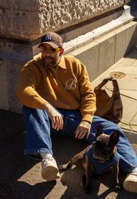 Une personne portant un pull moutarde avec un logo "B" est assise par terre à côté d'un bouledogue français noir portant un harnais bleu.