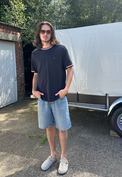 Camisa de manga corta navy con detalles en rojo y blanco, combinada con pantalones cortos de mezclilla azul claro y zuecos grises, posada al aire libre.