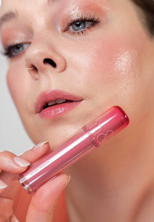 Frau mit rosa Lidschatten und glänzenden Lippen hält rosa Lipgloss nahe am Gesicht und zeigt die Details und Textur des Make-ups.