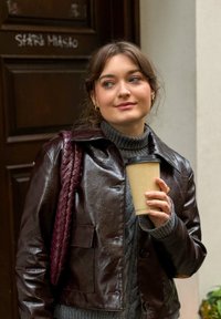 Bruine leren jas met een gestructureerde afwerking, grijze geribbelde trui, gevlochten bordeauxrode tas, met een beige koffiekop in de hand. Achtergrond van een donkerhouten deur.