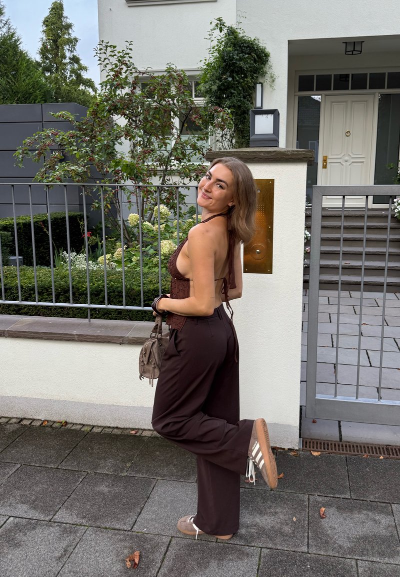 Rjav halter top, ohlapne temno rjave široke hlače, bež čevlji z belimi črtami, drži majhno torbico, stoji blizu moderne hiše in zelenja.