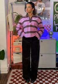 Femme portant un haut à manches longues Nike rayé rose et brun et un pantalon noir, debout les mains dans les poches dans une pièce avec des étagères et des affiches.