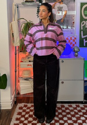 Mujer con una camiseta de manga larga Nike de rayas rosas y marrones y pantalones negros, de pie con las manos en los bolsillos en una habitación con estanterías y pósters.