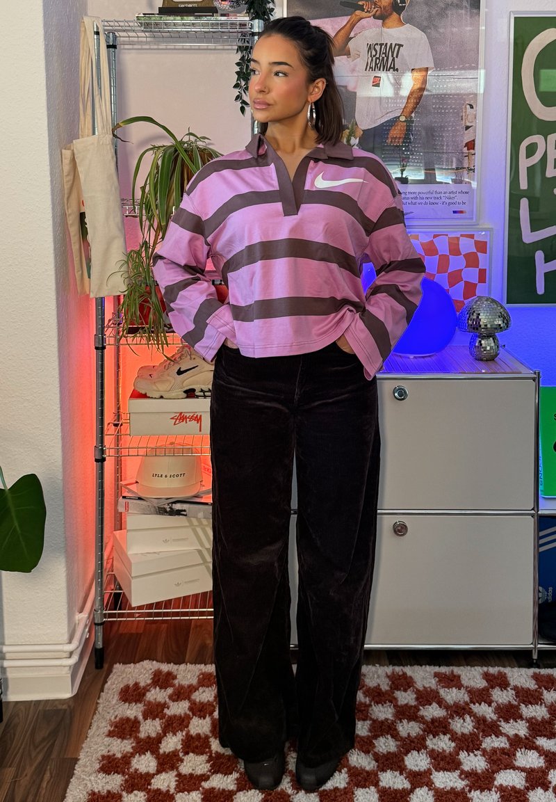 Femme portant un haut à manches longues Nike rayé rose et marron et un pantalon noir, debout les mains dans les poches dans une pièce avec des étagères et des affiches.