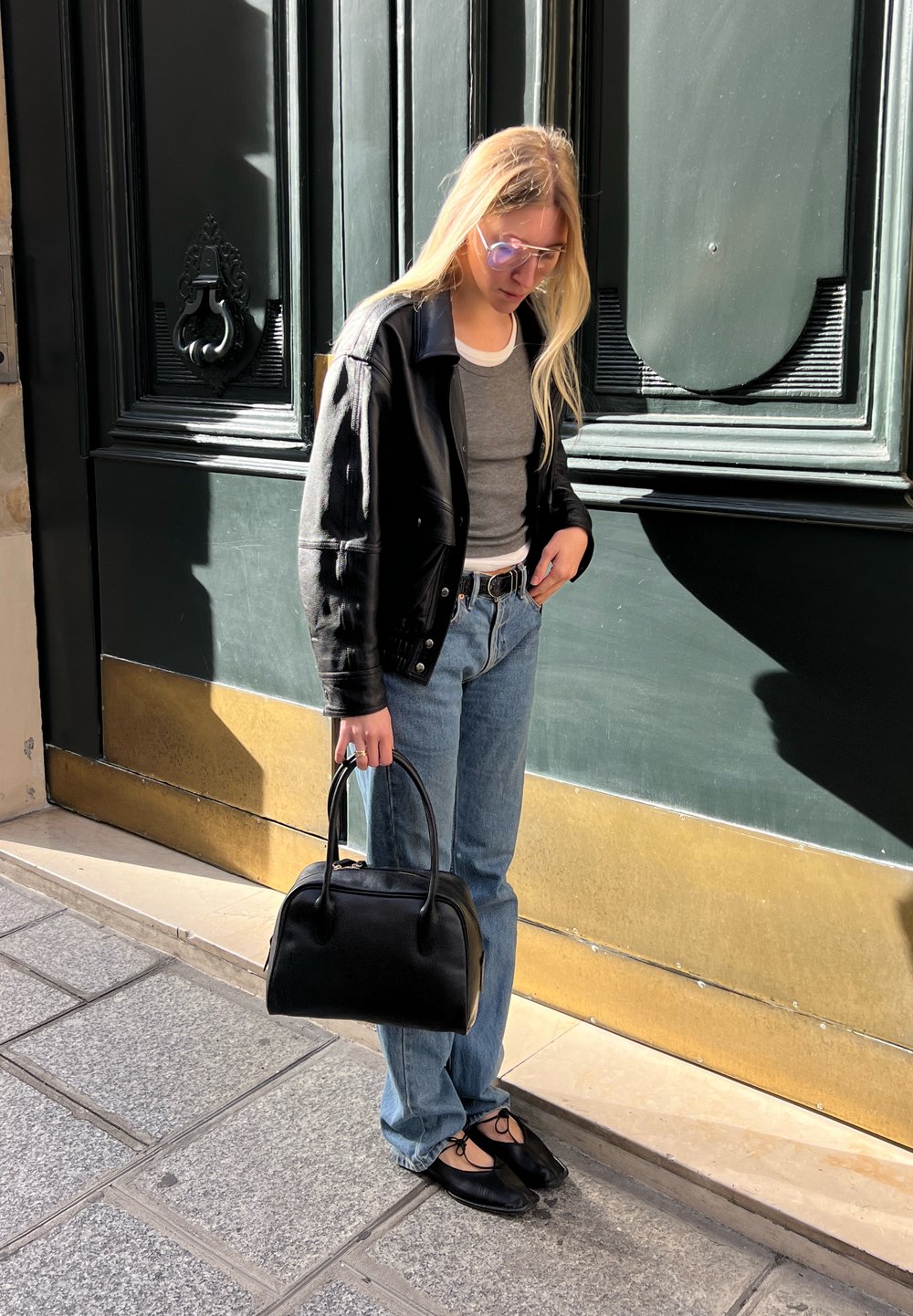 Crna kožna jakna preko sive majice, plave farmerke, crne balerinke, drži malu crnu torbicu; sunčane naočale s laganim ljubičastim nijansama; stoji na otvorenom.