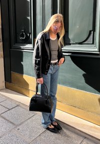 Crna kožna jakna preko sive majice, plave farmerke, crne balerinke, drži malu crnu torbicu; sunčane naočale s laganim ljubičastim nijansama; stoji na otvorenom.