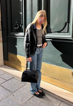Chaqueta de cuero negra sobre una camiseta gris, jeans azules, zapatos planos negros, sosteniendo un pequeño bolso negro; gafas de sol con tinte lila claro; de pie al aire libre.
