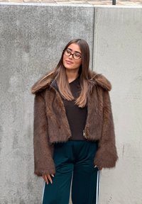 Brun kort jacka i konstpäls med huva, dragkedja och vida ärmar. Bärs över en mörk topp och gröna träningsbyxor med vita ränder.