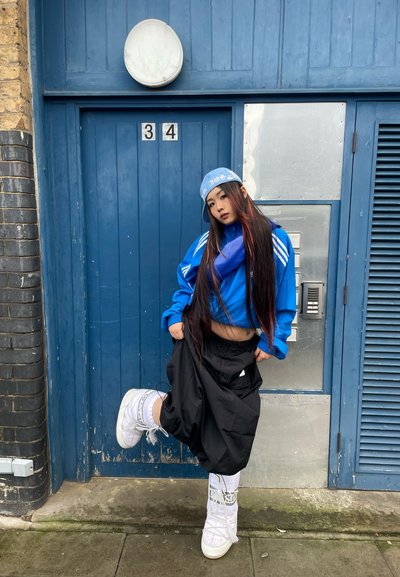 Chaqueta azul con cremallera y rayas blancas, pantalones holgados negros y botas blancas. Cabello largo y oscuro con reflejos rojos. Gorra azul en la cabeza.