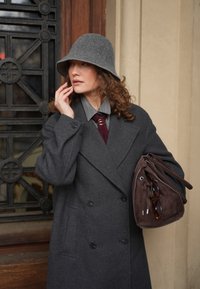 Femme en manteau gris et chapeau tenant un sac marron avec des lunettes de soleil, touchant son visage, debout près d'une porte en métal noire ornée et d'un mur beige.