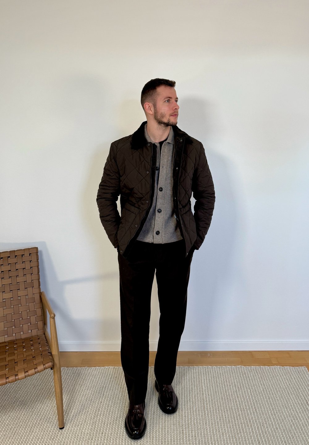 Homme debout à l'intérieur portant une veste matelassée marron, un pull gris boutonné, un pantalon noir et des mocassins foncés, près d'une chaise en bois tressé.