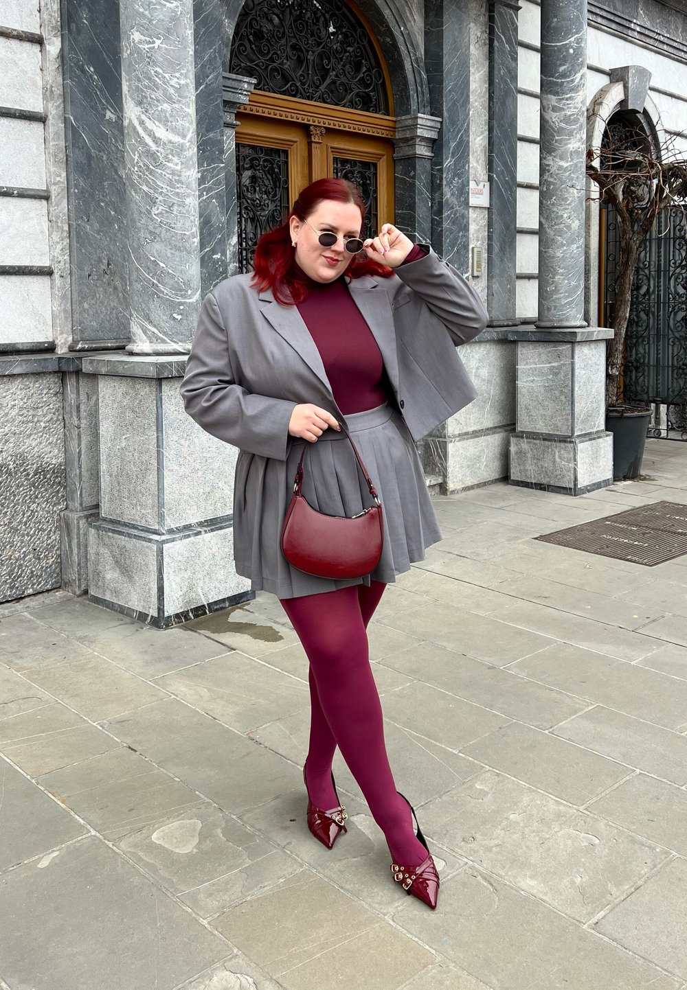 Γκρίζα σακακιού κομμένη πάνω από μπορντό τοπ και πτυχωτή φούστα. Συμβατά μπορντό καλσόν και καφέ ψηλοτάκουνα παπούτσια με διακοσμητικές λεπτομέρειες. Μικρή κόκκινη τσάντα.