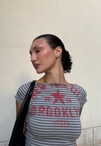 Harmaa ja valkoinen raidallinen t-paita, jossa on punainen graafinen teksti "NEW YORK CITY BROOKLYN 2009" ja tähtikuvio. Mallilla on siisti, sitottu hiustyyli.