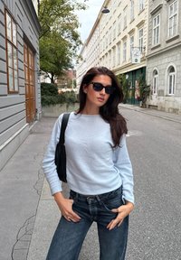 Maglione azzurro chiaro con scollatura a girocollo, abbinato a jeans blu scuro. Borsa a spalla nera e occhiali da sole completano l'outfit. Sfondo urbano.