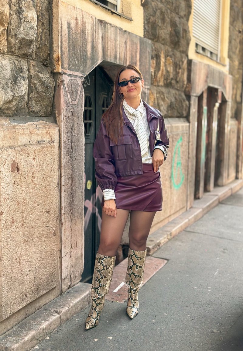 Femme portant des lunettes de soleil, une veste et une jupe en cuir violettes, un haut rayé et des bottes à motif serpent, debout sur un trottoir de ville près d’un mur en pierre.