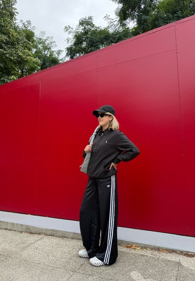 Chaqueta negra con logo, pantalones a rayas negros, sandalias blancas y bolsa gris, frente a una pared roja vibrante con vegetación arriba.
