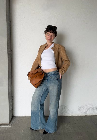 Mujer con chaqueta marrón claro, top corto blanco, jeans de pierna ancha, zapatos negros, sosteniendo un gran bolso marrón, de pie frente a una pared blanca.