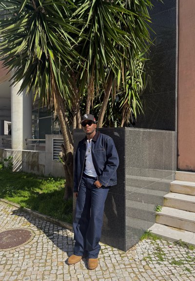 Hombre con chaqueta oscura, camisa blanca, pantalones oscuros, sombrero y gafas de sol apoyado contra una estructura de piedra negra cerca de una palmera al aire libre.