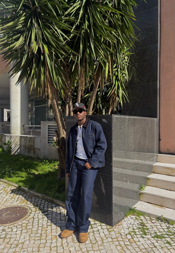 Mann mit dunkler Jacke, weißem Hemd, dunkler Hose, Hut und Sonnenbrille lehnt sich an eine schwarze Steinstruktur in der Nähe einer Palme im Freien.