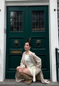En grøn dobbeltstående dør med løvehovedklopper. En person i en beige trenchcoat, hvid skjorte og lys nederdel knæler på trappen.