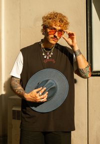 Schwarzes ärmelloses Shirt über einem weißen T-Shirt, blauer Strohhut, orangefarbene Sonnenbrille, geschichtete Perlenhalsketten und Tattoos auf den Armen.