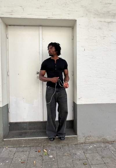 Hombre vestido con una camiseta negra de manga corta acanalada y jeans de pierna ancha color gris oscuro, sosteniendo auriculares, de pie sobre un escalón de piedra junto a una pared blanca.