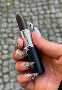 MAC-lippenstift in een zwarte huls met zilverkleurige accenten; rijke donkerbruine kleur; hand met lange, sierlijke nagels met zilveren accenten houdt de huls vast.