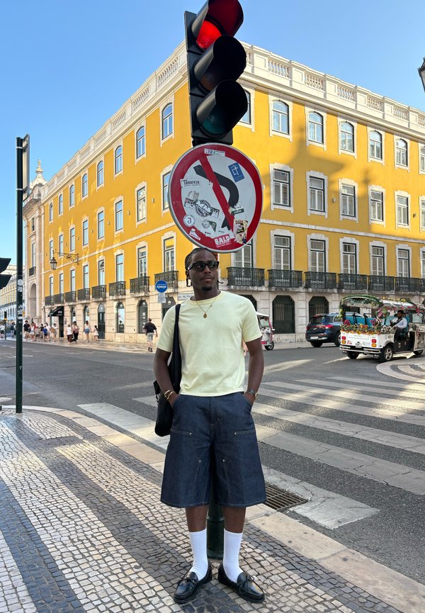 Uomo con occhiali da sole, camicia giallo chiaro, pantaloncini di jeans in piedi su un attraversamento pedonale vicino a un semaforo rosso e un edificio giallo.