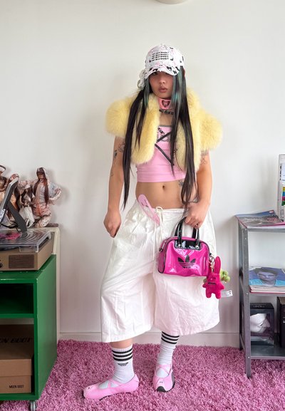 Persona joven con cabello largo lleva un crop top rosa, shrug de piel amarillo, pantalones blancos anchos, gorra rosa, sostiene un pequeño bolso rosa con llaveros de peluche.