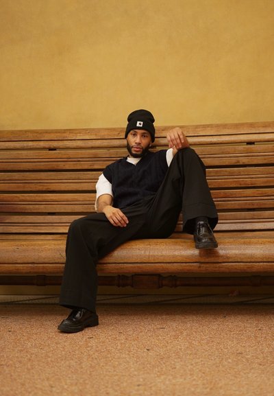 Gorro negro, chaleco de punto en color azul marino sobre una camisa blanca, pantalones anchos negros y zapatos negros, sentado en un banco de madera contra una pared texturizada.