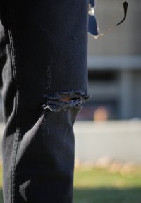 Black denim jeans featuring a distressed design with several frays and a prominent hole at the knee. Smooth texture and tapered fit.