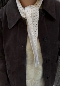 Brown suede jacket with a collar and black buttons, layered over a cream knit sweater. A textured white scarf is tied at the neck.