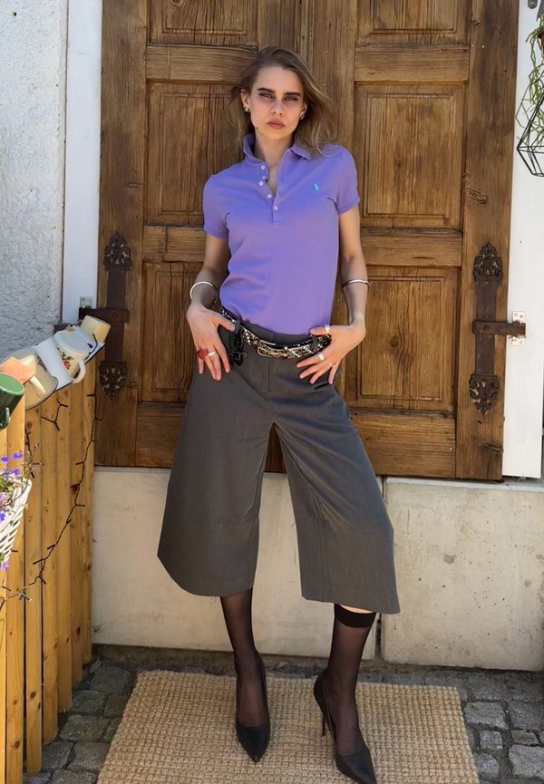Joven mujer vistiendo una camiseta tipo polo morada, pantalones grises de pierna ancha, medias negras transparentes y tacones negros, de pie frente a una puerta de madera.