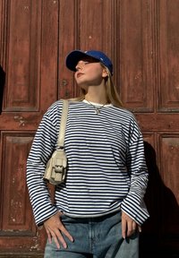 Rööpüline pikade varrukatega särk valge ja tumesinise värviga, heledad denim-jeans ja metallik ristkott. Taustal on tekstuuriga punased puidust uksed.