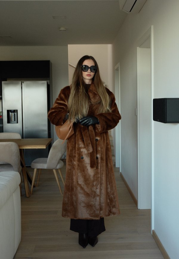 Femeie purtând o haină maro de blană, mănuși negre, ochelari de soare și ținând un geamantan maro, stând într-o bucătărie modernă cu zonă de luat masa.