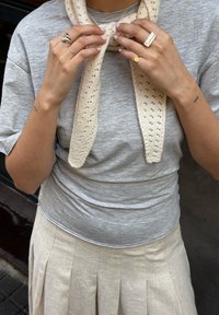 Harmaa lyhythihainen paita, johon on yhdistetty vaalea beige neulehuivi, jossa on pitsimäinen kuvio. Käsissä, joissa on minimalistisia koruja, on sormuksia.