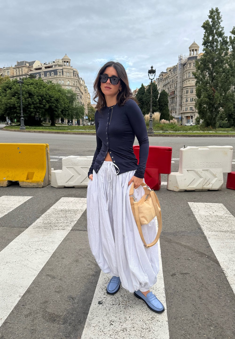Navy long-sleeve top with buttons, white striped wide-leg trousers, blue shoes, and a tan handbag. Pedestrian area with barriers in the background.