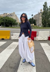 Navy long-sleeve top with buttons, white striped wide-leg trousers, blue shoes, and a tan handbag. Pedestrian area with barriers in the background.