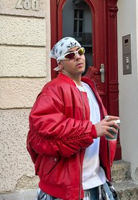 Červená kožená bomber jacket s staženými rukávy, bílý tričko, šedé kostkované kalhoty, bílý šátek a sluneční brýle. Drží kávový hrníček.