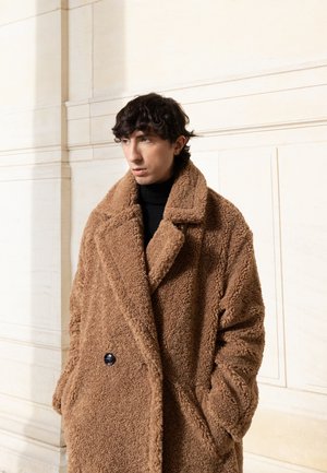 Brun oversized shearlingjakke med en dyb krave, tekstureret stof og en knapdetalje. Båret over en sort turtleneck i et veloplyst miljø.