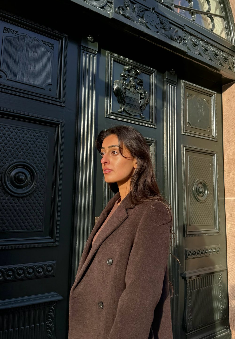 Καφέ παλτό διπλής όψης με υφή, φορεμένο πάνω από ένα ανοιχτόχρωμο τοπ. Σκοτεινή διακοσμημένη πόρτα στο φόντο με περίπλοκες λεπτομέρειες και υλικά.