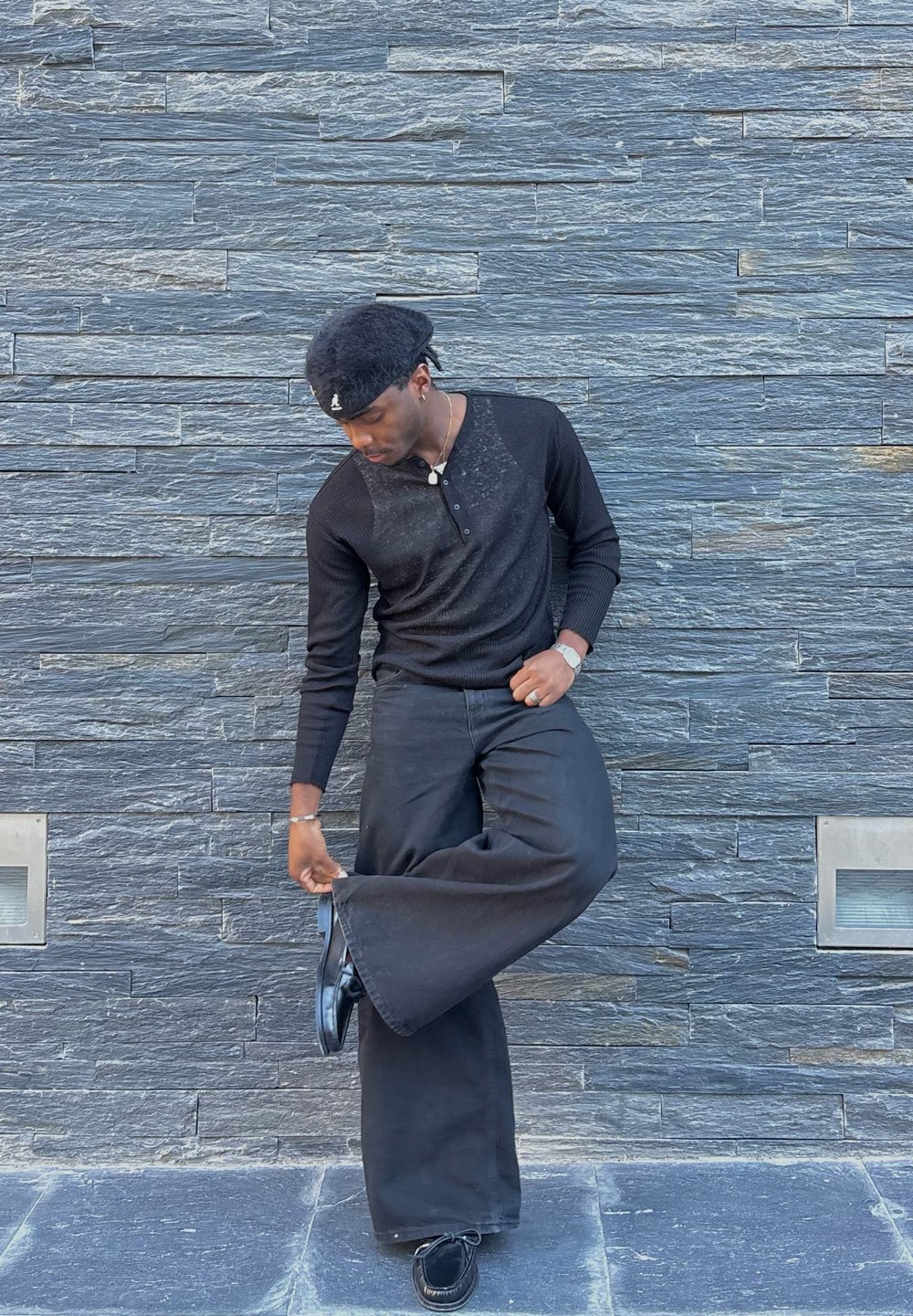 Camisa henley negra de manga larga, pantalones de pierna ancha negros y zapatos negros brillantes. El fondo presenta una pared de piedra gris texturizada.