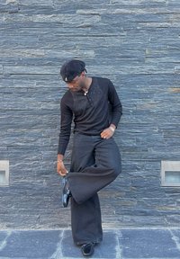 Camisa henley negra de manga larga, pantalones de pierna ancha negros y zapatos negros brillantes. El fondo presenta una pared de piedra gris texturizada.